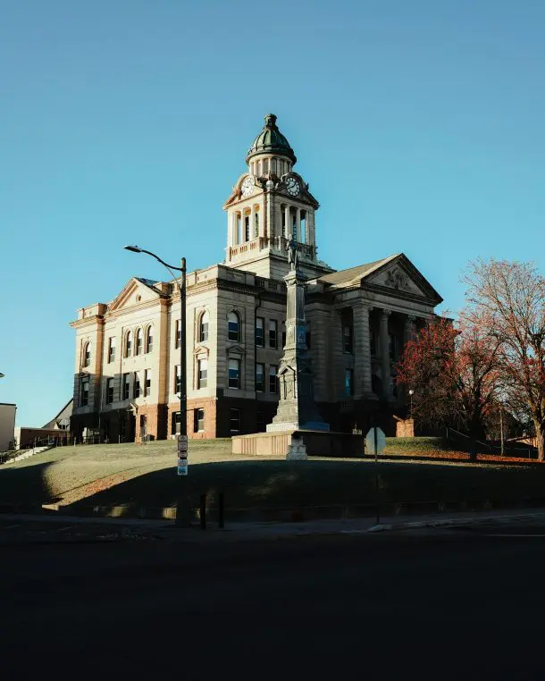 Neoclassical courthouse stands atop a grassy hill, its clock tower rising above a stone monument in front, bathed in warm light against a clear blue sky. No legible text.
