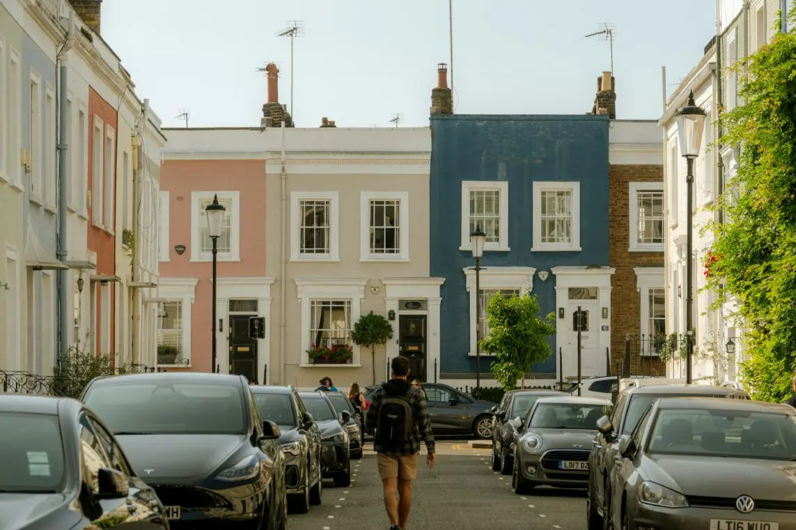 A person walking down a quiet residential street lined with parked cars, facing a row of colourful terraced houses in pastel pink, cream, blue and brick, with small trees and classic street lamps.