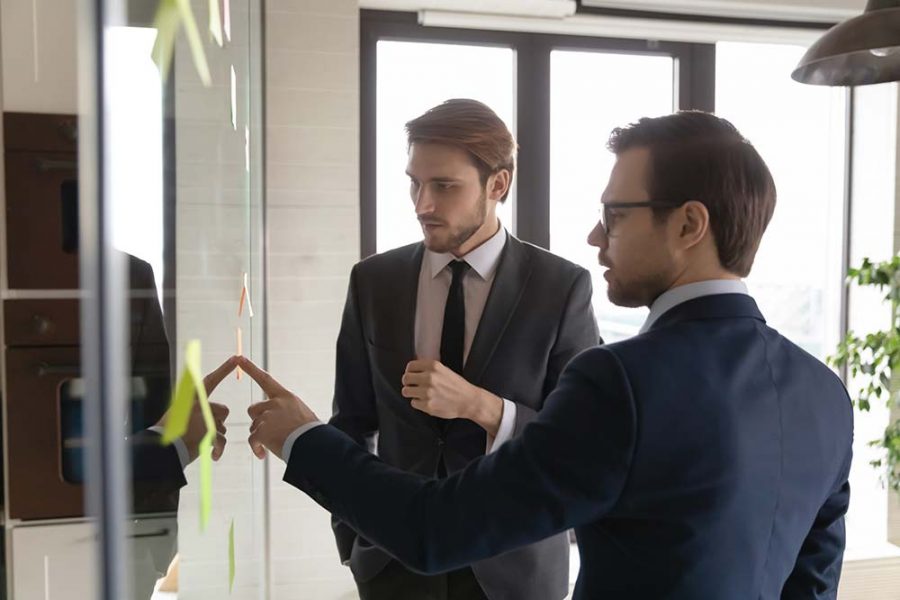 Two men work together at a white board, the man closest to the camera is pointing to the board